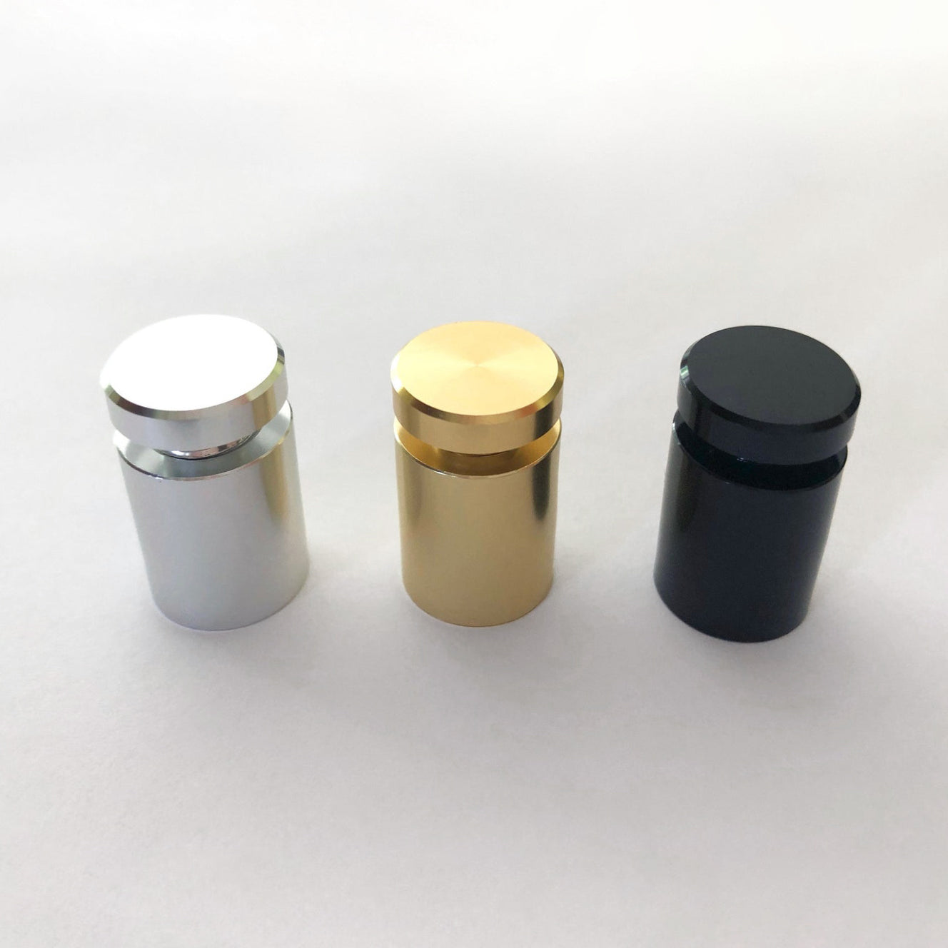 Three metallic cylindrical standoffs in silver, gold, and black support the Large Acrylic Calendar with Bottom Notes from Girl Friday Shop on a white surface. Each has a screw top and smooth finish, ideal for wet-erase markers.