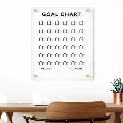 A Goal Chart | Sales Tracker for Real Estate by Girl Friday, displaying customizable house icons under Homes Sold and Sales Volume, hangs as a floating acrylic board above the wooden desk, accompanied by a chair, a small plant, and a copper mug near the window.
