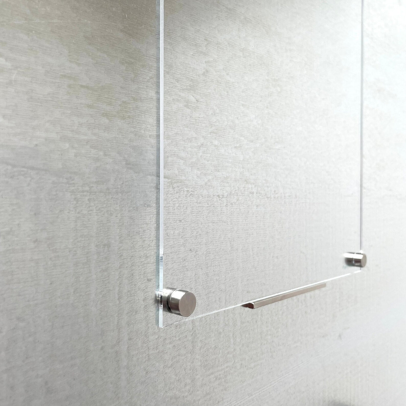 A close-up of a glass panel secured by sleek, cylindrical Girl Friday Bar magnets against a textured wall. The minimalist design is enhanced with a hint of gold, elevating its modern appeal. The subtle pattern on the wall complements the refined aesthetic perfectly.