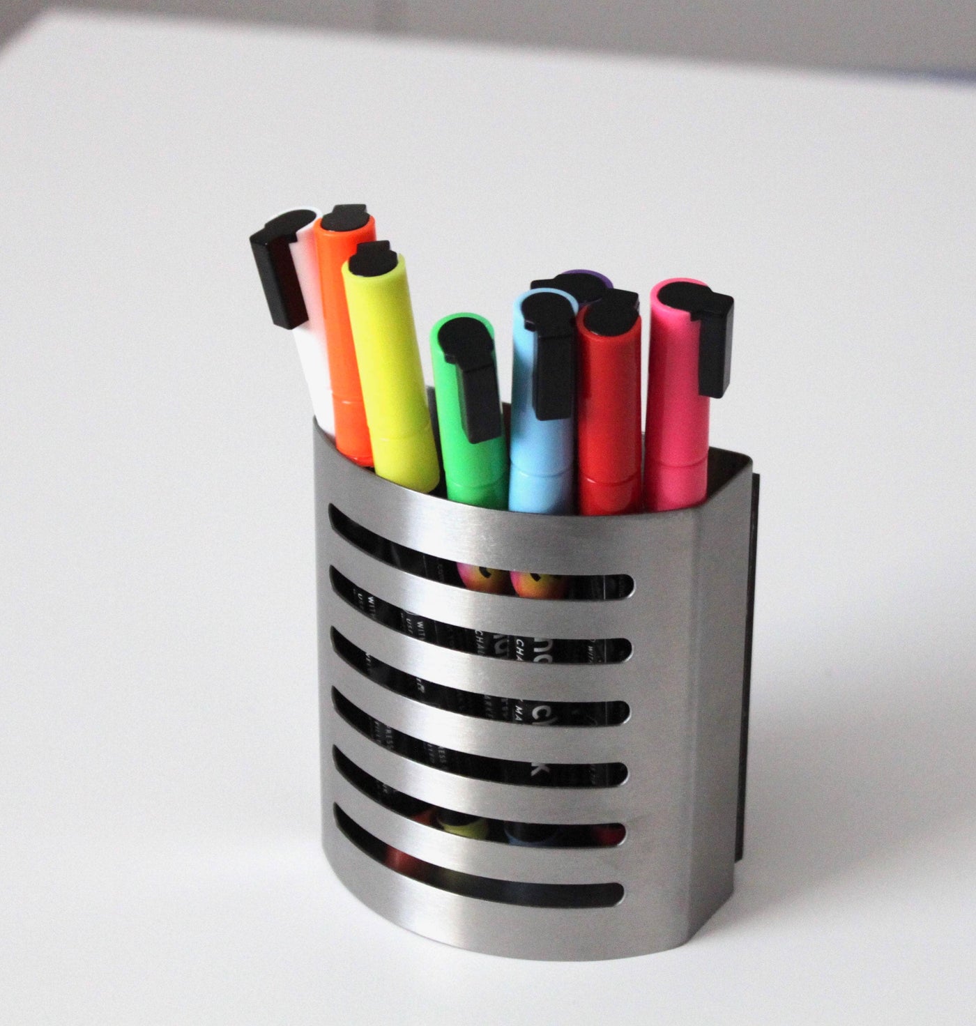 A sleek, slotted stainless steel cup holder rests on a white surface, showcasing Girl Fridays Chalk Ink Vibrant Markers in black, orange, green, blue, pink, red, and yellow.