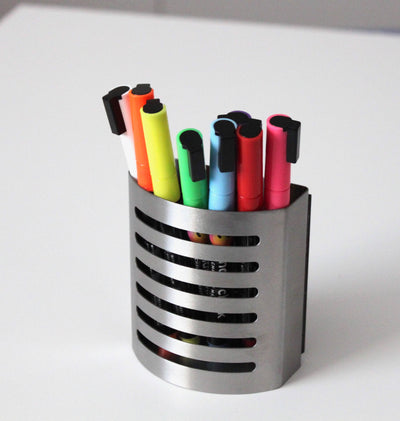 A sleek, slotted stainless steel cup holder rests on a white surface, showcasing Girl Fridays Chalk Ink Vibrant Markers in black, orange, green, blue, pink, red, and yellow.