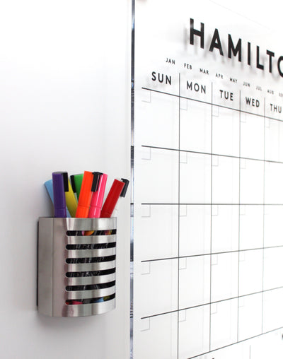 A colorful wall-mounted holder contains Girl Fridays Chalk Ink Vibrant Markers next to a large dry-erase calendar with months, weekdays, and a task grid.