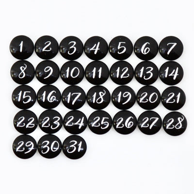 Black circular magnets numbered 1-31 in white script are arranged on a grid pattern against the premium CalendarGirlCompanys 24 x 18 Framed Whiteboard Calendar.