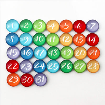 The CalendarGirlCompanys 24 X 18 Framed Whiteboard Calendar (#18203) features colorful glass stones numbered 1 to 31 on a white surface, with rows transitioning from blue to purple, creating a vibrant monthly calendar.