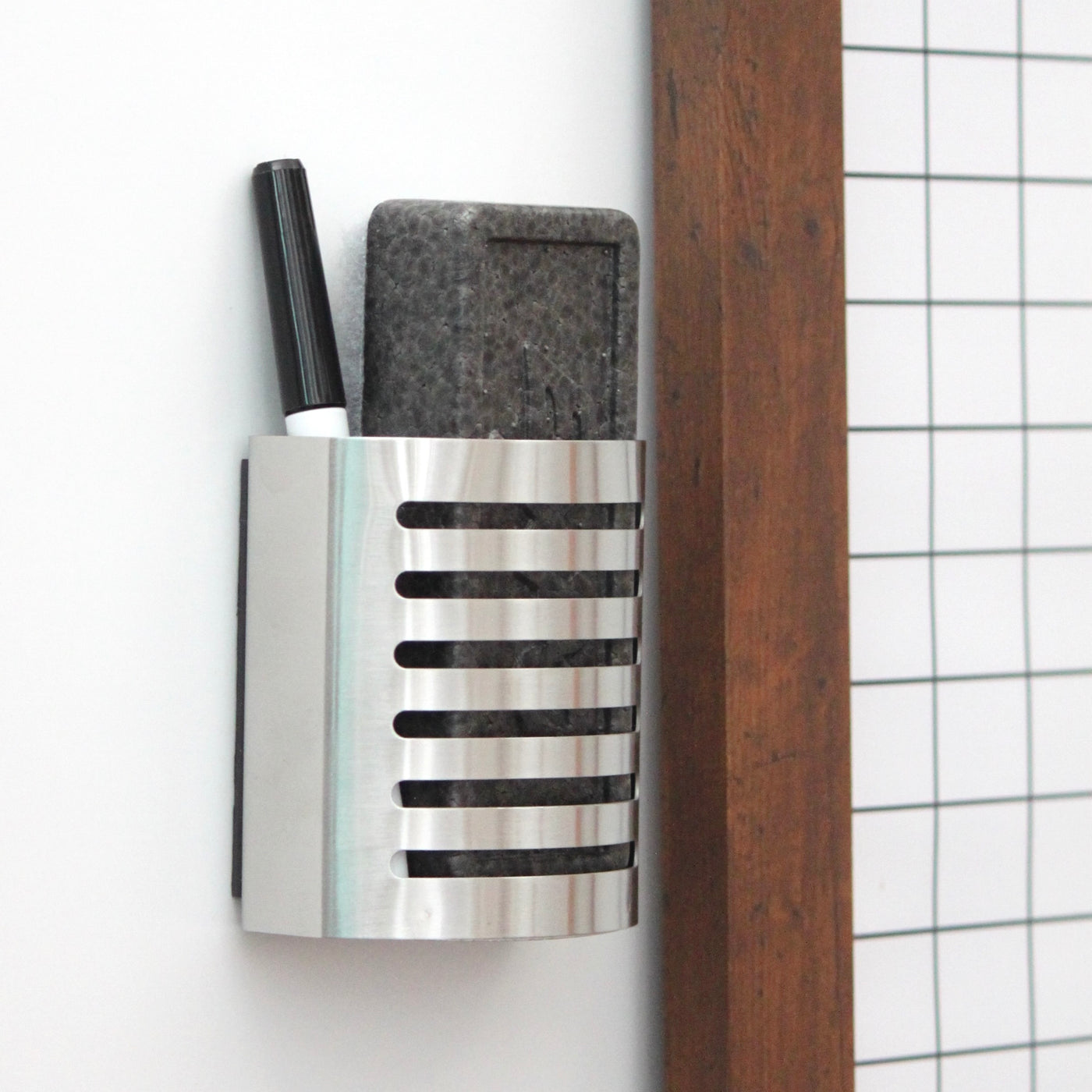 A Girl Friday stainless steel marker holder is mounted on a white wall next to a wooden grid-framed structure. It contains a black pen and an EXPO eraser and features horizontal slits.