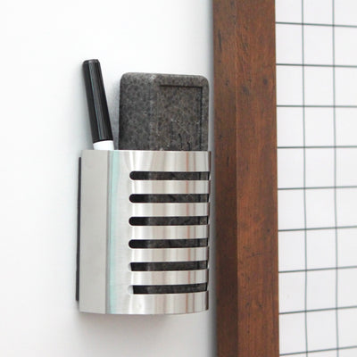 A wall-mounted metallic holder with vertical slits is attached to the premium acrylic whiteboard and holds a black marker and gray eraser. Positioned next to Girl Friday Shops 18x24 framed In/Out Magnetic Staff Board, it combines functionality with style. Multi-colored magnets included.