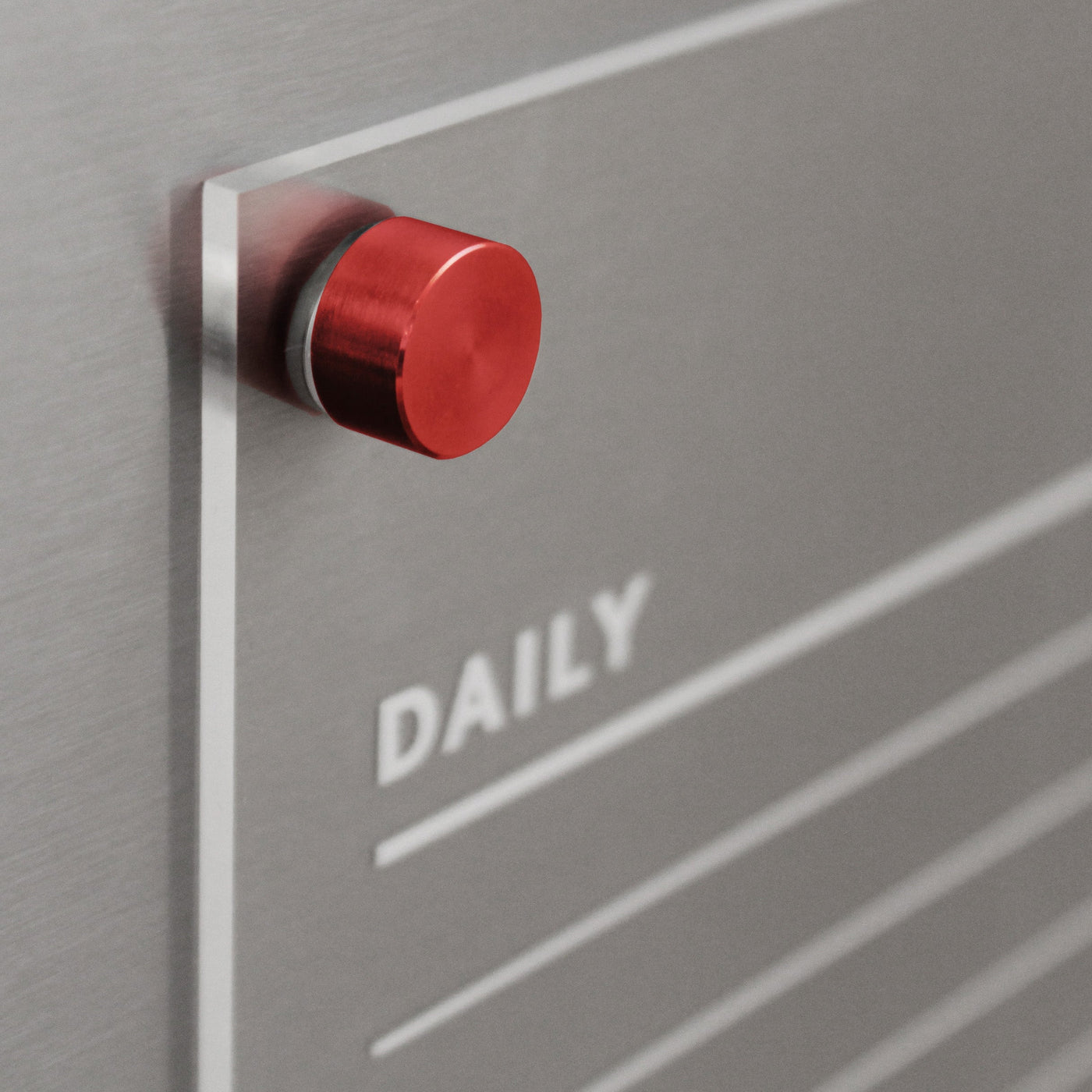 A sleek metallic surface features a red cylindrical knob on the left. DAILY is printed in white, with thin lines below for wet-erase notes, making it ideal as a magnetic board. Its the Acrylic Fridge Chore Chart for 2 children by Girl Friday, perfect for an efficient fridge organizer.