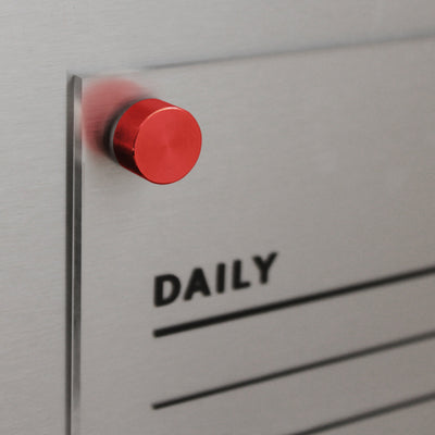 Close-up of a red circular magnet on Girl Fridays Acrylic Fridge Chore Chart for 2 children, set against a metallic board with a transparent sheet. The board reads DAILY with lines, ideal for planning or scheduling and perfect for updates using liquid chalk ink markers.