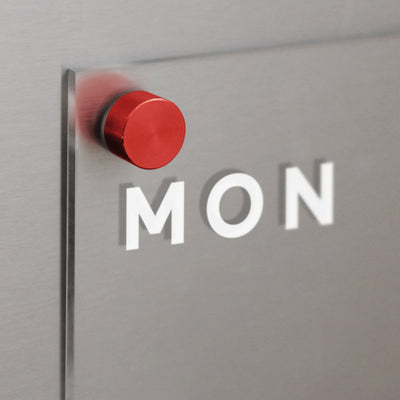 The Girl Friday Clear Acrylic Weekly Fridge Calendar, featuring white text and an embossed MON, is secured to a metallic surface with a red magnetic pin, casting soft shadows.