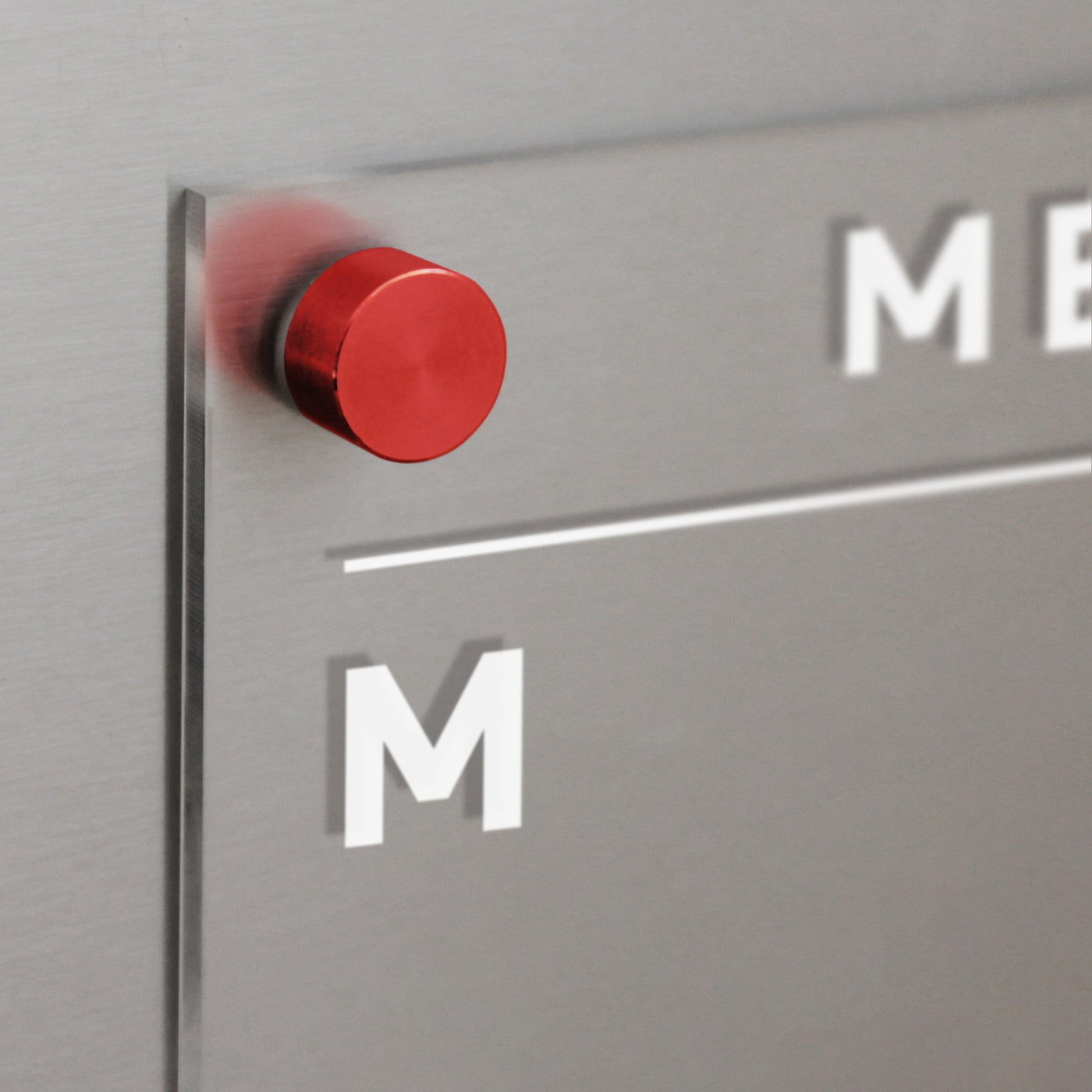 A close-up of a silver metallic surface with the Girl Friday Acrylic Fridge Menu Board. The letter M is visible on the board, and a red cylindrical magnet slightly blurs its surroundings, creating an ideal space for organizing with dry-erase markers.