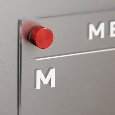 A close-up of a silver metallic surface with the Girl Friday Acrylic Fridge Menu Board. The letter M is visible on the board, and a red cylindrical magnet slightly blurs its surroundings, creating an ideal space for organizing with dry-erase markers.