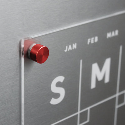 Close-up of the Girl Friday Shops Clear Acrylic Monthly Fridge Calendar with a magnetic surface, displaying S and M. The months JAN, FEB, and MAR appear at the top, while a red magnetic pin secures notes. Ideal for organizing with wet-erase markers.