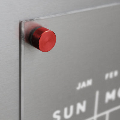 Close-up of a red magnet on Girl Fridays Acrylic Fridge Calendar, wall-mounted and perfect for organizing your week. The sleek display features days in white text against a metallic background. Use wet-erase markers to personalize it with your custom family name.