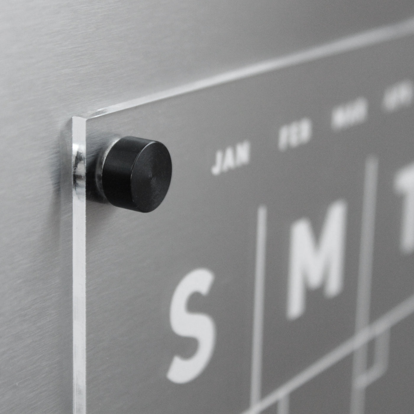 A close-up of Girl Fridays Clear Acrylic Monthly Fridge Calendar shows a sleek, wall-mounted design with partially visible weekdays. A black knob holds a transparent sheet for JAN and FEB. Ideal for wet-erase markers, it has a brushed metal background and optional custom family name.
