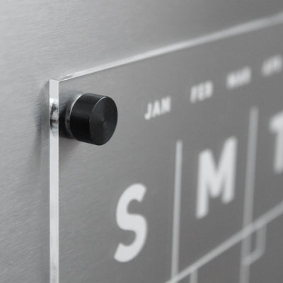 A close-up of Girl Fridays Clear Acrylic Monthly Fridge Calendar shows a sleek, wall-mounted design with partially visible weekdays. A black knob holds a transparent sheet for JAN and FEB. Ideal for wet-erase markers, it has a brushed metal background and optional custom family name.
