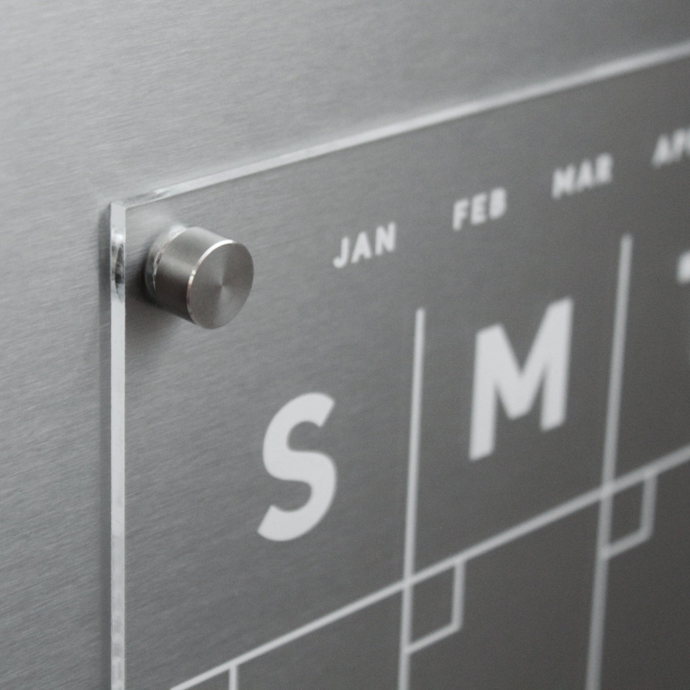 A close-up showcases a metallic perpetual wall calendar paired with Girl Fridays Clear Acrylic Monthly Fridge Calendar, featuring custom family names and labeled days S and M. The minimalist design is modern, with months above and enhanced by a sleek circular side knob.