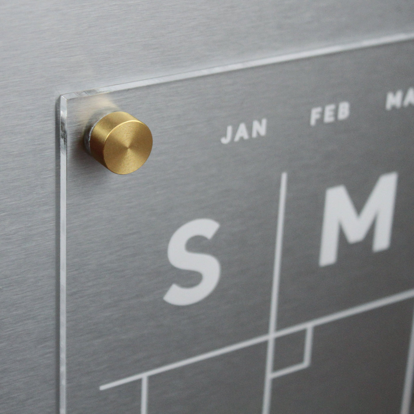 Close-up of the Girl Friday Shops Clear Acrylic Monthly Magnetic Fridge Calendar with magnetic fasteners. White text shows weekdays, while JAN, FEB, and MAR are at the top. Ideal for wet-erase markers, this calendar elegantly mounts on any metal surface.