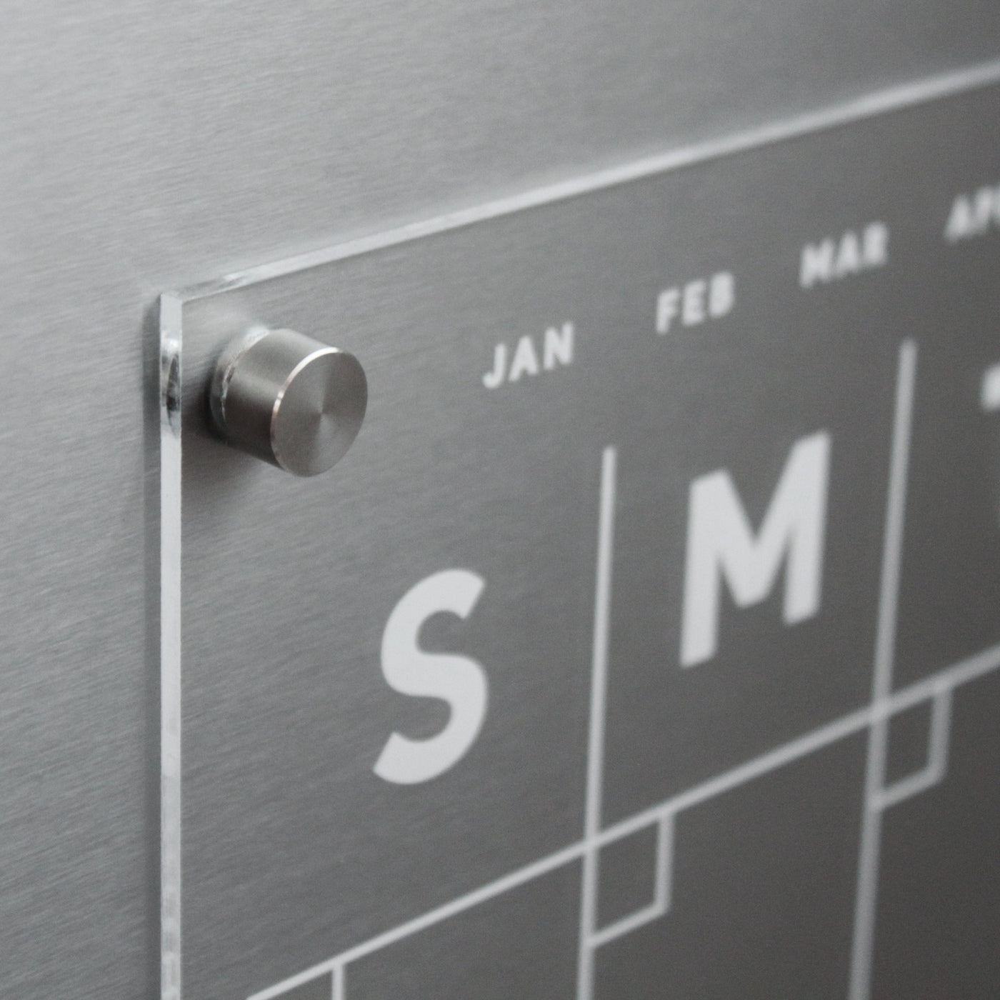 The Clear Acrylic Monthly Magnetic Fridge Calendar by Girl Friday Shop features sleek metallic accents, bold white S and M for days, JAN, FEB, and MAR for months. Mounted with a silver pin on magnets, it includes a wet-erase marker for easy updates.