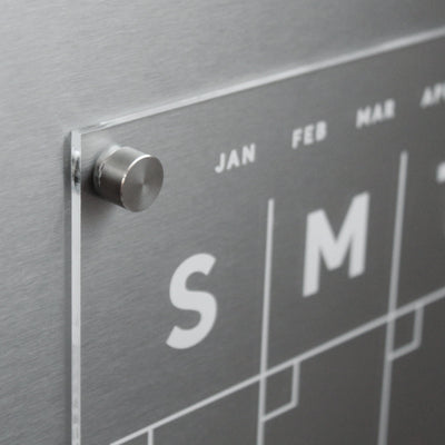 The Clear Acrylic Monthly Magnetic Fridge Calendar by Girl Friday Shop features sleek metallic accents, bold white S and M for days, JAN, FEB, and MAR for months. Mounted with a silver pin on magnets, it includes a wet-erase marker for easy updates.