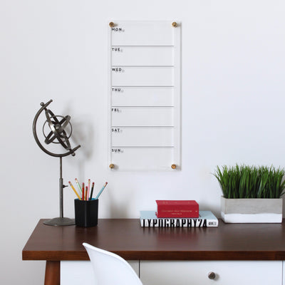 A minimalist office setup features a wooden desk with a globe sculpture, pencil holder, red and white books, and a white chair. A Girl Friday Shop Clear Acrylic Weekly Wall Calendar with gold accents mounts on the wall, compatible with wet-erase markers. Nearby is a green plant in a white pot.