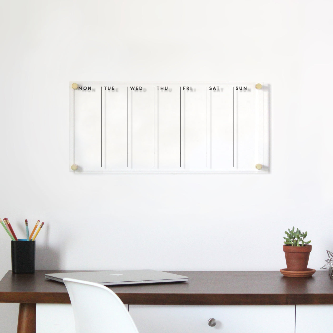 A minimalist workspace includes a wooden desk with a laptop, an acrylic organizer filled with colorful pencils, and a small potted plant. Above the desk is Girl Friday Shops Acrylic Calendar-weekly planner for wet-erase markers, labeled Monday to Sunday.