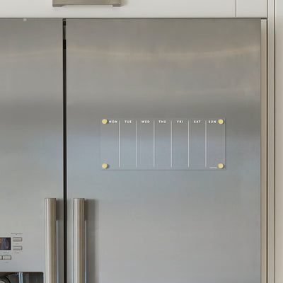 The Clear Acrylic Weekly Fridge Calendar by Girl Friday features a stainless steel-friendly design with white text. It displays columns for each day starting Monday and is held up by yellow magnets. Compatible with wet-erase markers, its perfect for easy adjustments throughout the week.
