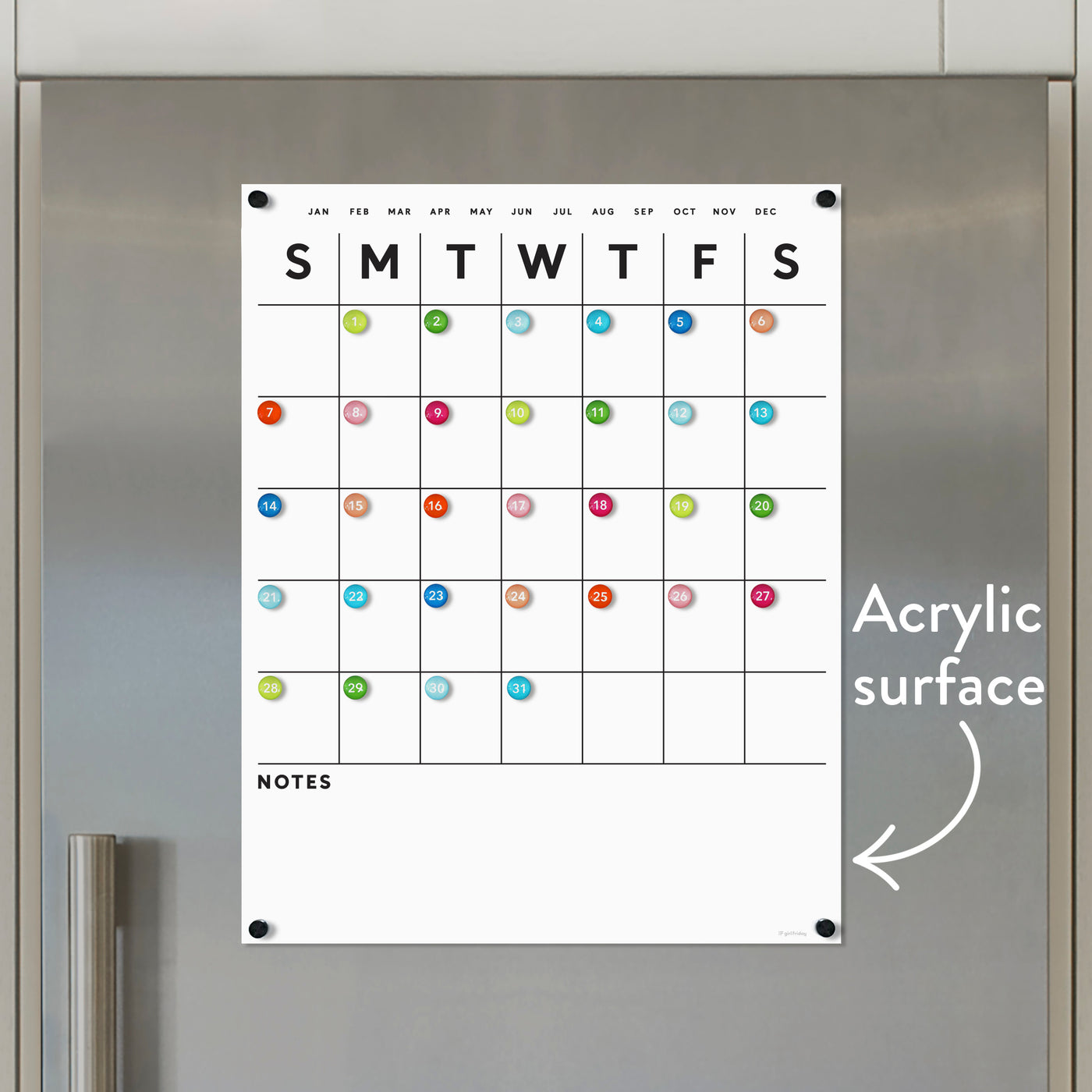 The White Acrylic Fridge Calendar by Girl Friday Shop features days, months, and a multicolored numbered grid. Below is a Notes section, with an arrow indicating the dry erase surface. Neodymium magnets secure it to a metallic background.