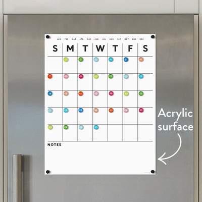 The White Acrylic Fridge Calendar by Girl Friday Shop features days, months, and a multicolored numbered grid. Below is a Notes section, with an arrow indicating the dry erase surface. Neodymium magnets secure it to a metallic background.