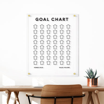 A Goal Chart | Sales Tracker for Real Estate by Girl Friday, featuring customizable houses labeled Homes Sold and Sales Volume, hangs above a desk adorned with a wooden chair and potted plant, creating an inviting workspace.