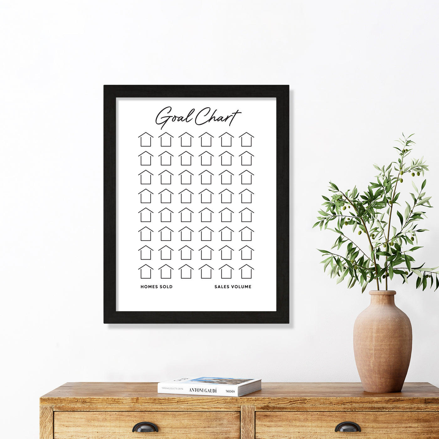 A Girl Friday Shop Framed Goal Chart | Sales Tracker for Real Estate, made from premium acrylic with a house motif, adorns a white wall above a wooden table. The table features stacked books and a beige vase filled with green branches.