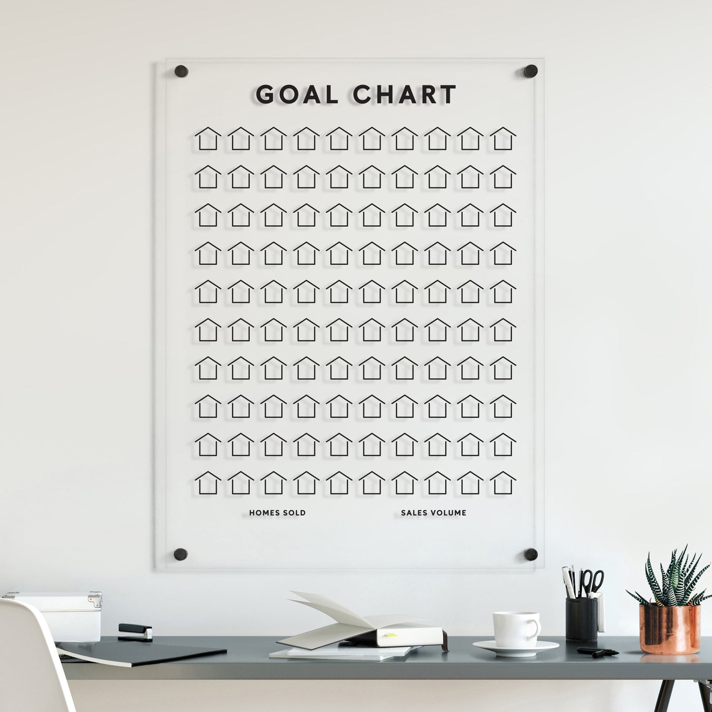 Above the desk, Girl Fridays Goal Chart | Sales Tracker for Real Estate elegantly displays rows of customizable houses tracking Homes Sold and Sales Volume. The desk holds a laptop, an open notebook, a pen cup, and a small plant in a copper pot.