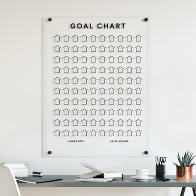 Above the desk, Girl Fridays Goal Chart | Sales Tracker for Real Estate elegantly displays rows of customizable houses tracking Homes Sold and Sales Volume. The desk holds a laptop, an open notebook, a pen cup, and a small plant in a copper pot.