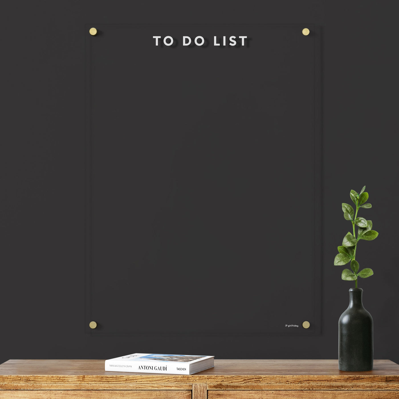 The Girl Friday Shops Clear Acrylic Customizable Memo Board with white text stands vertically as a To Do List on a dark wall, joined by a small plant in a black vase and two books on wood. Its smooth surface awaits wet-erase markers, leaving the task area invitingly empty.