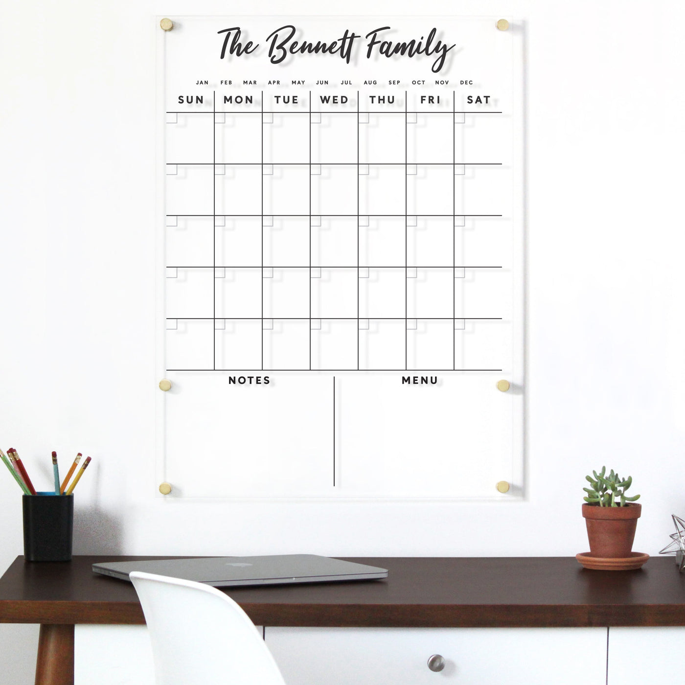 The Girl Friday Shops Clear Acrylic Customizable Monthly Wall Calendar with Script Font features a sleek, wall-mounted design titled The Bennett Family, perfect for organizing days, notes, and menu plans. The high-quality hardware enhances its modern aesthetic alongside a desk with a plant and pencils.