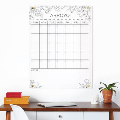 The Clear Acrylic Monthly Wall Calendar by Girl Friday Shop features floral designs with your family name, displayed professionally. It hangs above a wooden desk with a laptop, books, a potted plant, and a silver geometric ornament. Four rows of empty boxes await your plans.