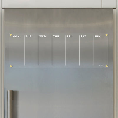 On a metal fridge door, the Girl Friday Clear Acrylic Weekly Fridge Calendar with white text is displayed horizontally. The calendar, featuring Monday through Sunday columns and attached with four yellow magnets, is blank—ideal for jotting down notes using wet-erase markers.
