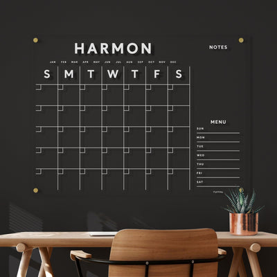 The Clear Acrylic Customizable Monthly Wall Calendar by Girl Friday Shop features labeled weekdays, a notes section, and a menu. Its mounted on a dark wall over a wooden desk with a chair. A potted plant decorates the space, while its ghosting-free cleaning ensures it stays pristine.