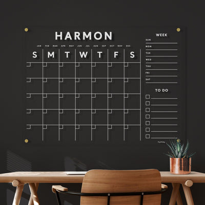 The Harmon black wall-mounted planner elegantly complements the décor, featuring a grid layout with days of the week and to-do list lines. The ghosting-free design ensures easy cleaning, enhancing your workspace style with a subtle touch above your wooden desk and chair ensemble.