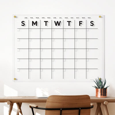 The Clear Acrylic Monthly Wall Calendar from Girl Friday Shop showcases a minimalist design with daily squares and days of the week at the top. Below, a wooden desk enhances the setting with a potted succulent in sunlight, while wet-erase markers stand ready for your plans.