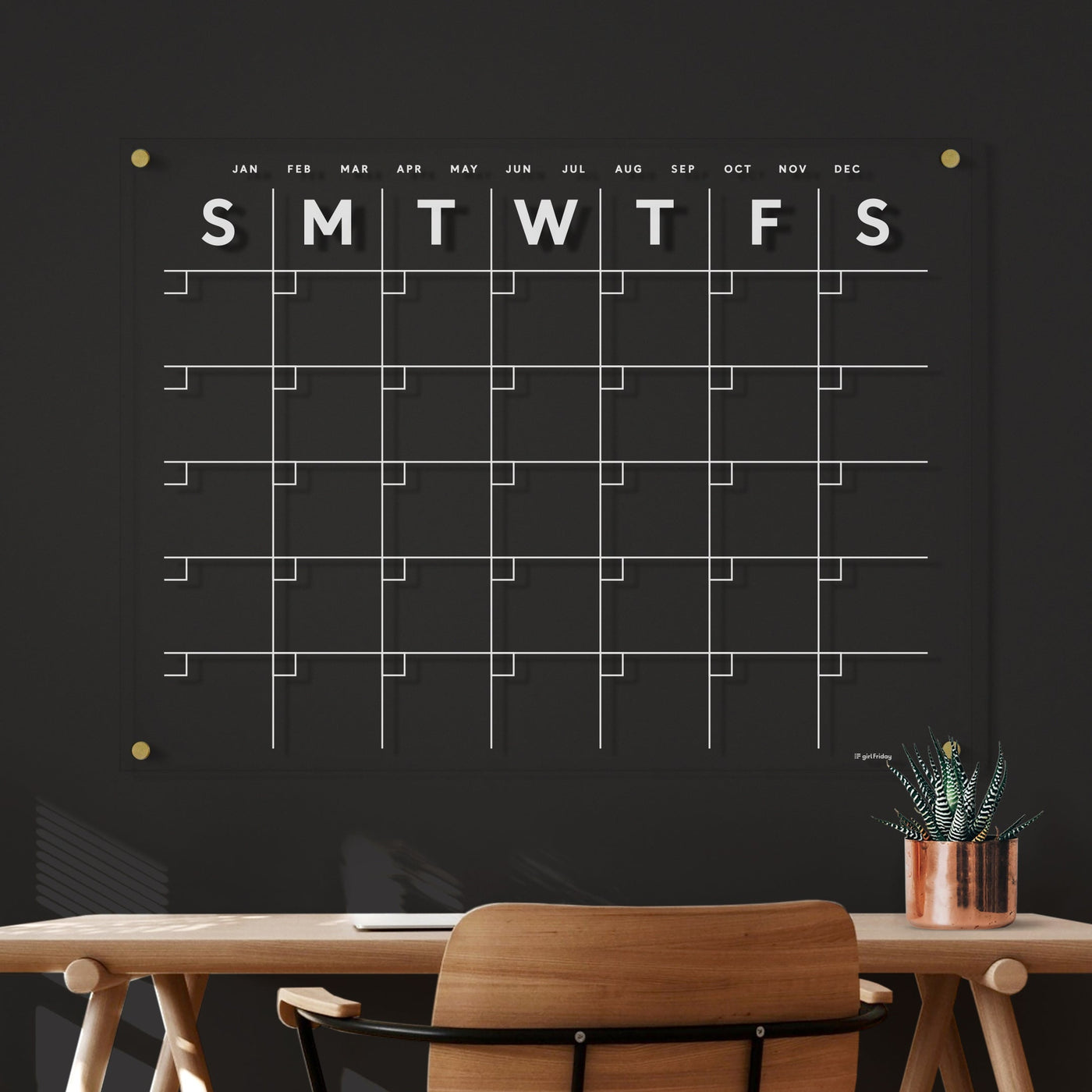 The Clear Acrylic Monthly Wall Calendar by Girl Friday Shop, featuring white text on a sleek black surface, hangs elegantly above a wooden desk with a potted plant. Its horizontal design and modern finish complement the dark gray wall perfectly, displaying days from Sunday to Saturday.