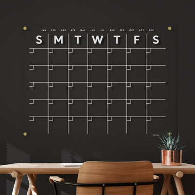 The Clear Acrylic Monthly Wall Calendar by Girl Friday Shop, featuring white text on a sleek black surface, hangs elegantly above a wooden desk with a potted plant. Its horizontal design and modern finish complement the dark gray wall perfectly, displaying days from Sunday to Saturday.