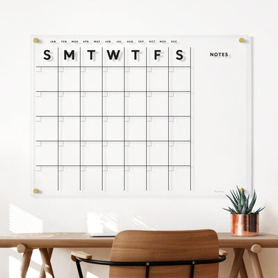 The Clear Acrylic Monthly Wall Calendar with Notes by Girl Friday Shop, featuring columns for each day of the week and a notes section, hangs above a wooden desk. A small potted plant accessorizes the desk, while wet-erase markers are neatly placed nearby.