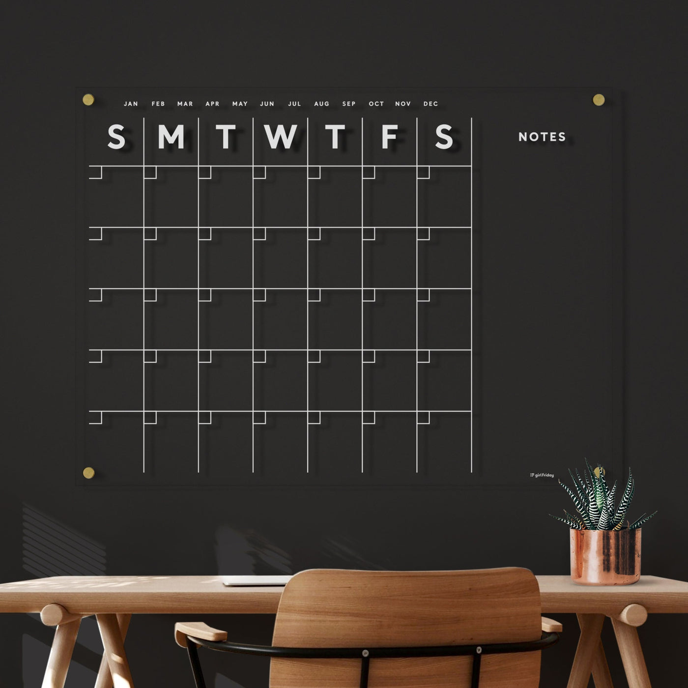 A black acrylic Acrylic Calendar with side notes by Girl Friday Shop showcases a weekly grid for notes with wet-erase markers. It hangs above a wooden desk and chair, while natural light casts shadows on a copper potted plant, adding charm to the space.