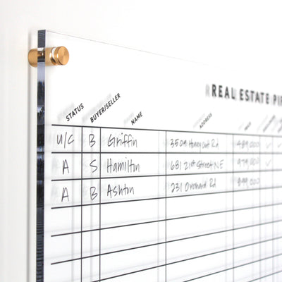 A close-up of a Girl Friday Real Estate Pipeline Acrylic Board on the wall, shimmering with gold pegs. The clear surface displays columns for status, buyer/seller, name, and address in wet-erase marker with entries like Griffin, Hamilton, and Ashton.