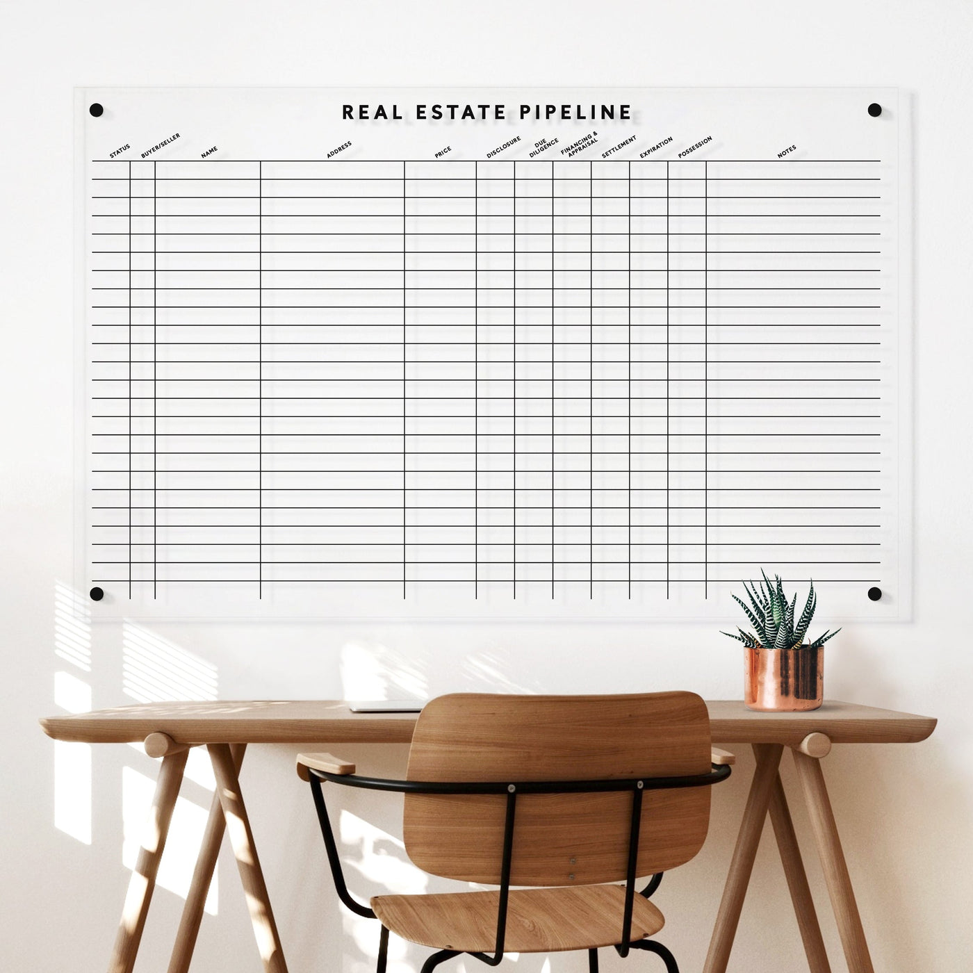 A wooden desk with a chair is set against a white wall. Above it is the Girl Friday Real Estate Pipeline acrylic board for tracking properties. A small plant in a copper pot adorns the desk next to wet-erase markers ready for updates.