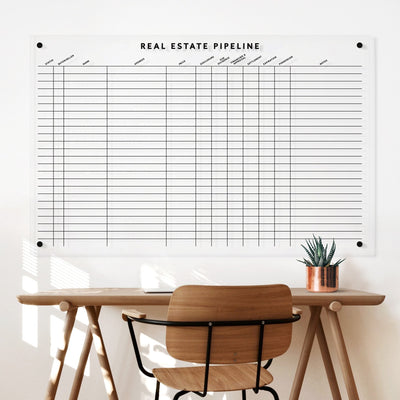 A wooden desk with a chair is set against a white wall. Above it is the Girl Friday Real Estate Pipeline acrylic board for tracking properties. A small plant in a copper pot adorns the desk next to wet-erase markers ready for updates.