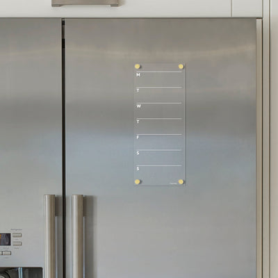 The Clear Acrylic Weekly Fridge Calendar by Girl Friday, with white text, attaches to a stainless steel refrigerator using yellow magnets for each day of the week (M, T, W, T, F, S, S), and pairs perfectly with wet-erase markers for easy planning.