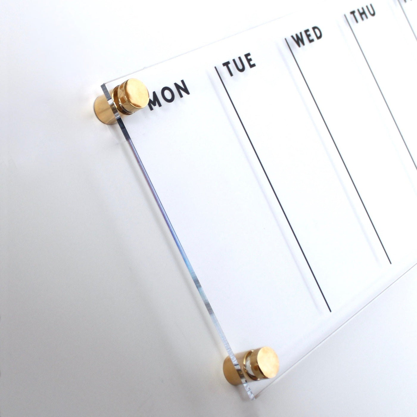 The Clear Acrylic Weekly Wall Calendar | Horizontal from Girl Friday Shop features a sleek design with gold mounts and a glossy acrylic surface, perfect for wet-erase markers. It includes bold black letters for each weekday, starting with Monday.