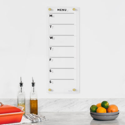 The Girl Friday Clear Acrylic Customizable Weekly Menu Board features initials for each day and lines for meals. Below, a counter holds fruit, tall corked bottles, and a lidded red casserole dish. Ideal for wet-erase markers.