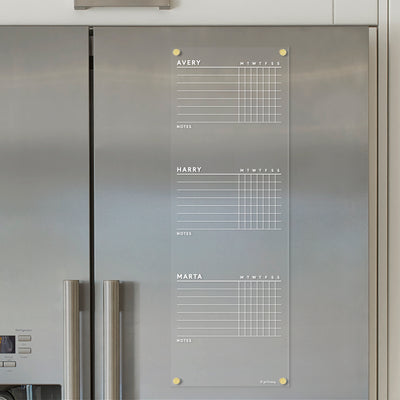 A Girl Friday Acrylic Fridge Chore Chart for 3, 4, or 5 children is attached to a stainless steel refrigerator. It features vibrant liquid chalk ink markers with sections for Avery, Harry, and Marta, complete with days-of-the-week checkboxes and a notes area.